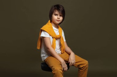 Yakışıklı, modacı, 11-13 yaşlarında. Stüdyoda yeşil arka planda bir sandalyede oturuyor. Sıradan bir tarz. Stüdyoda şık bir çekim.