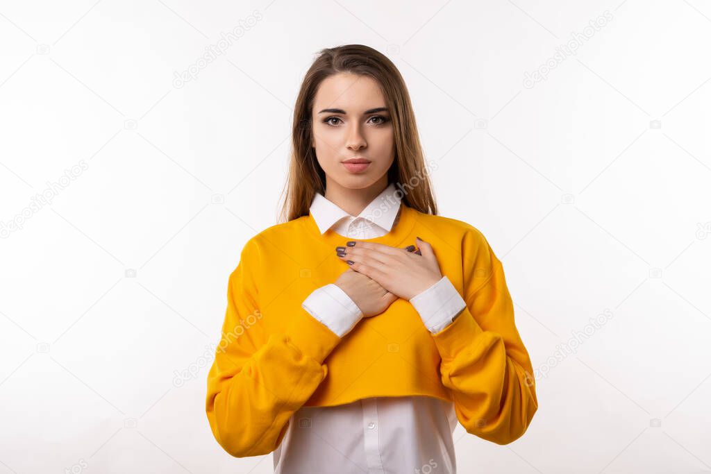Pleasant young brunette woman in casual clothes hold hands on heart cheerfully thanking for compliments or praises, feel grateful and pleased. Studio shot, white background