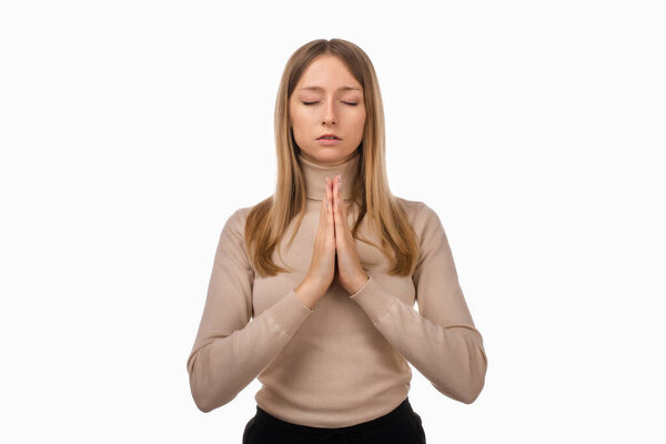 Attractive blond girl in casual clothes prays for wellness of family, keeps palms pressed together in praying gesture, saying namaste. Human emotions, gesture concept. Studio shot white background