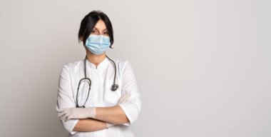 Koru kendini. Koruyucu yüz maskesi takan kadın doktor ya da bilim adamı, beyaz önlük ve steteskop gri arka planda çapraz kollarla ayakta duruyor. Tıp, meslek ve sağlık konsepti