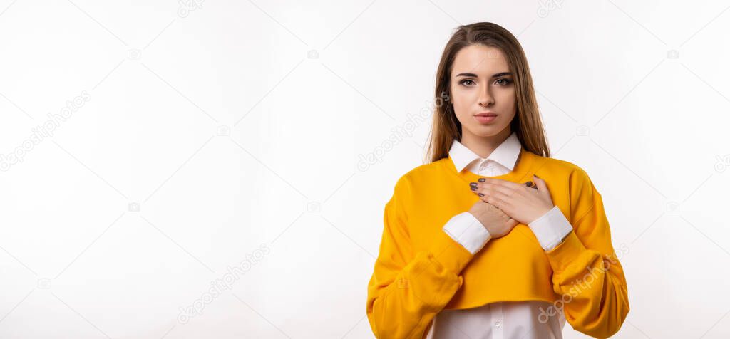 Pleasant young brunette woman in casual clothes hold hands on heart cheerfully thanking for compliments or praises, feel grateful and pleased. Studio shot, white background. Mock up copy space