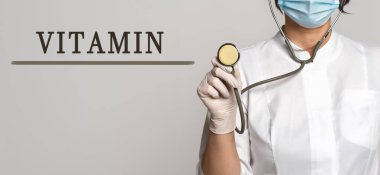 VITAMIN - gri arkaplanda metin kavramı. Yakınlarda beyaz önlüklü ve steteskoplu bir doktor var. Tıbbi konsept