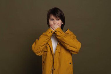 Günlük giysiler içinde korkmuş küçük bir çocuk ağzının yanında el ele tutuşuyor, korkudan titriyor, korkunç sesten çok korkuyor, yeşil arka planda sersemlemiş bir şekilde duruyor. Gerçek insan tepkileri, duyguları ve duyguları