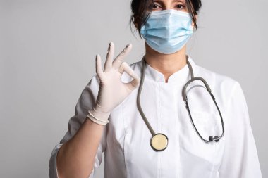 Koruyucu maske takan kadın doktor, laboratuvar önlüğü ve stetoskop tamam işareti gösteriyor. Koru kendini. Tıp ve sağlık hizmetleri kavramı