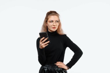 Kafası karışmış, sıradan giysiler içinde bir kenara bakıp akıllı telefonunu tutan güzel bir kadının görüntüsü. Teknoloji, gençlik ve iletişim konsepti. Beyaz arkaplan