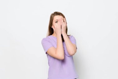 Endişeli, korkmuş, parmaklarının arasına bakan genç bir kadın, beyaz arka planda soyutlanmış. Yarım boy portre. İnsani duygular, yüz ifadesi kavramı
