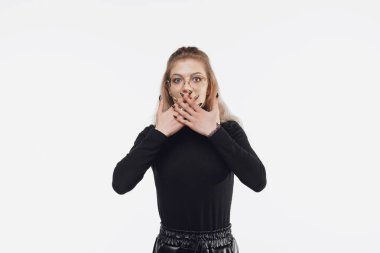 Tüh. Şok olmuş, siyah günlük kıyafetler giymiş genç bir kadın beyaz arka planda dikilen şok yüzünden ellerini ağzının üzerinde tutuyor.