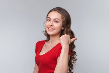 Neşeli genç esmer kadının portresi, baş parmağı ve gülümsemesi bir yana, insanları davet etmek tanıtım gibi görünüyor, gri arka planda duruyor.