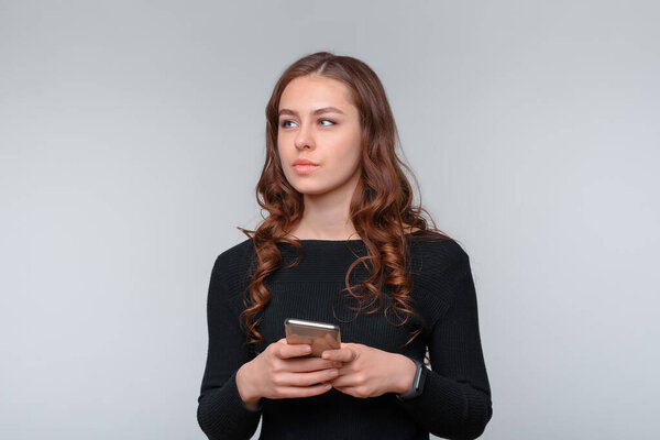 Portrait of nice young brunette woman holding smartphone, looking away over gray background