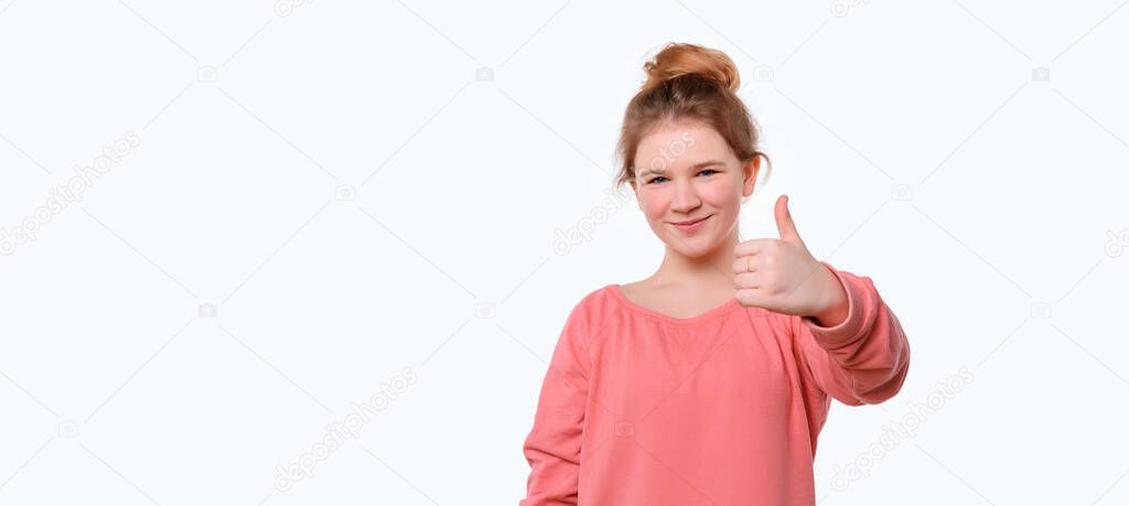 Portrait of a cute young girl in pink casual sweatshirt showing thumb up. White background. Human emotions, gesture concept