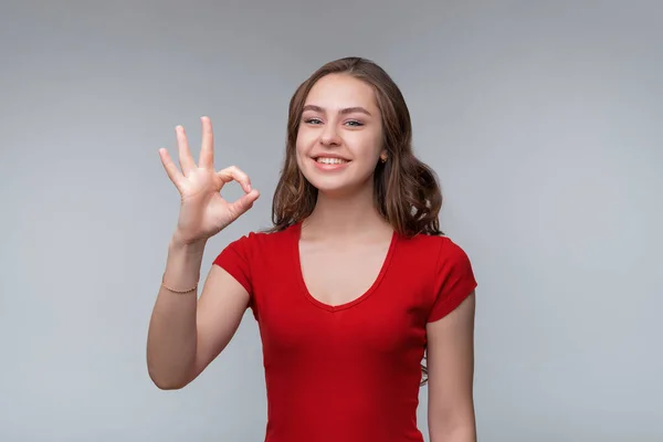 いい仕事だ 魅力的な若いブルネットの女性で赤TシャツショーOkサインと笑顔満足 肯定的な応答を与えます 良い同意サインを示します 大丈夫と承認にうなずきます 灰色の背景に立って — ストック写真