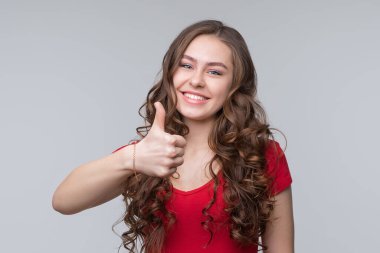 Uzun, kıvırcık saçlı, kırmızı tişörtlü güzel bir kadın baş parmağını kaldırıyor ve gülümsüyor. Kameraya bakan yüz ifadesi onaylanıyor