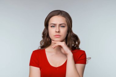 Hmm, ilginç. Düşünceli genç esmer kadın, kameraya bakıyor ve bir şeyler düşünüyor, varsayımda bulunuyor, düşünüyor. Stüdyo fotoğrafı, gri arka plan. İnsani duygular, yüz ifadesi kavramı