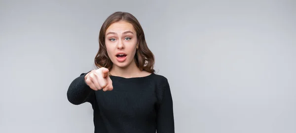 ItS your fault. Displeased young brunette woman is pointing finger at camera and to you, angry and furious with you. Studio shot, gray background. Human emotions, gesture concept