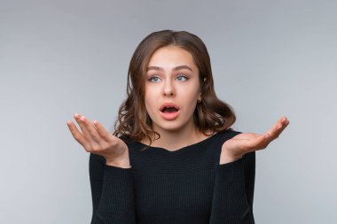 Kafası karışmış genç esmer kız öğrenci omuz silkiyor, ne yapacağını bilmiyor, bilgisiz ve gri arka planda şaşkınlık içinde duruyor. İnsani duygular, yüz ifadesi kavramı