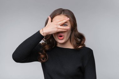 Korkmuş ve endişeli genç esmer kadının portresi, elleriyle gözlerini kapatıyor, parmaklarının arasından endişeli, paniklemiş, gri arka planda duruyor.