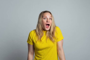 Şaşırmış ve heyecanlı olgun bir kadın kameraya şaşkın şaşkın bakıyor ve eğleniyor. Reklamlardan etkilenmiş, gri arka planda günlük sarı tişörtle ayakta duruyor.