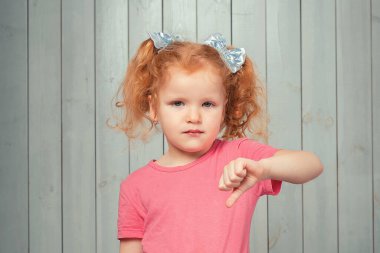 Ciddi kızıl saçlı, 4-6 yaşlarında pembe tişörtlü, baş parmağını aşağı gösteren ve hoşlanmadığını ifade eden bir kız. Stüdyo çekimi, hafif ahşap arka plan