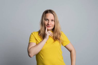 Confused mature woman wearing basic yellow t shirt looking at camera with pensive look. Studio shot, gray background