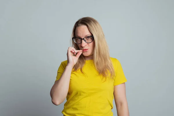 Shh its secret. Portrait of mature woman in glasses showing zip gesture as if shutting mouth on key, promises to keep secret, wearing casual yellow t shirt, gray background
