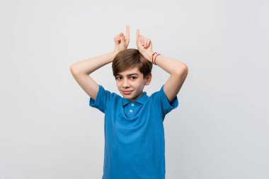 Pretty boy 12-14 years old in blue t shirt looking at the camera having fun, making silly faces and making horns with fingers. Studio shot, white background, isolated