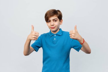 Güzel çocuk kameraya bakıyor ve açık gri arkaplanda baş parmak işareti yapıyor. 12-14 yaşlarındaki yakışıklı gencin portresi.