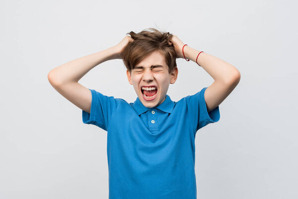 Frustrated boy with eyes closed, messing up and pulling his hair, hands to head shouting and screaming, isolated over light gray background. Stressed adolescent, failure concept