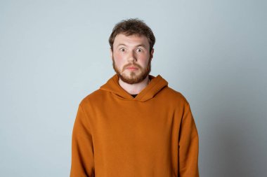 Portrait of shocked and scared bearded modern young man, looking amazed at something embarrassing, standing in stylish hoodie against studio gray background