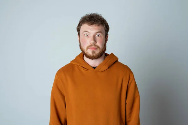 Portrait of shocked and scared bearded modern young man, looking amazed at something embarrassing, standing in stylish hoodie against studio gray background