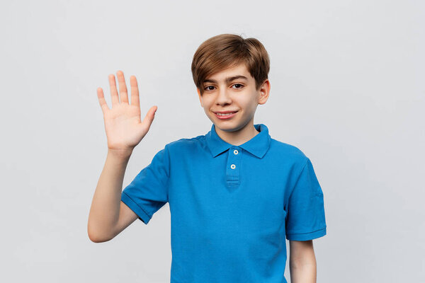 Красавчик, глядя на улыбающуюся камеру, здоровается с тобой на светло-сером фоне. Портрет красивого подростка 12-14 лет, машущего рукой