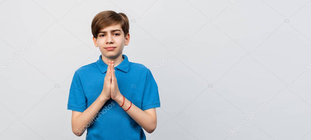 optimistic and hopeful teen boy praying with hands held together, waiting for results, awaiting miracle, praying with happy positive emotion, standing against light gray background