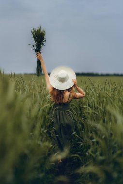 Şapkalı genç bir kadın buğday tarlasında yürüyor, hayattan ve yazdan zevk alıyor. Buğday tarlası. Güneş ışığı altında güzel doğa manzarası. Sağlıklı yaşam tarzı kavramı. Gürültülü görüntü, film tahılı