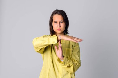 Portrait of beautiful teen girl making time out gesture, looking at camera, wearing casual yellow shirt, standing isolated on gray background clipart