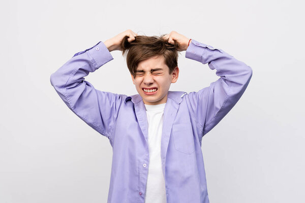 Frustrated boy with eyes closed, messing up and pulling his hair, hands to head shouting and screaming, isolated over light gray background. Stressed adolescent, failure concept