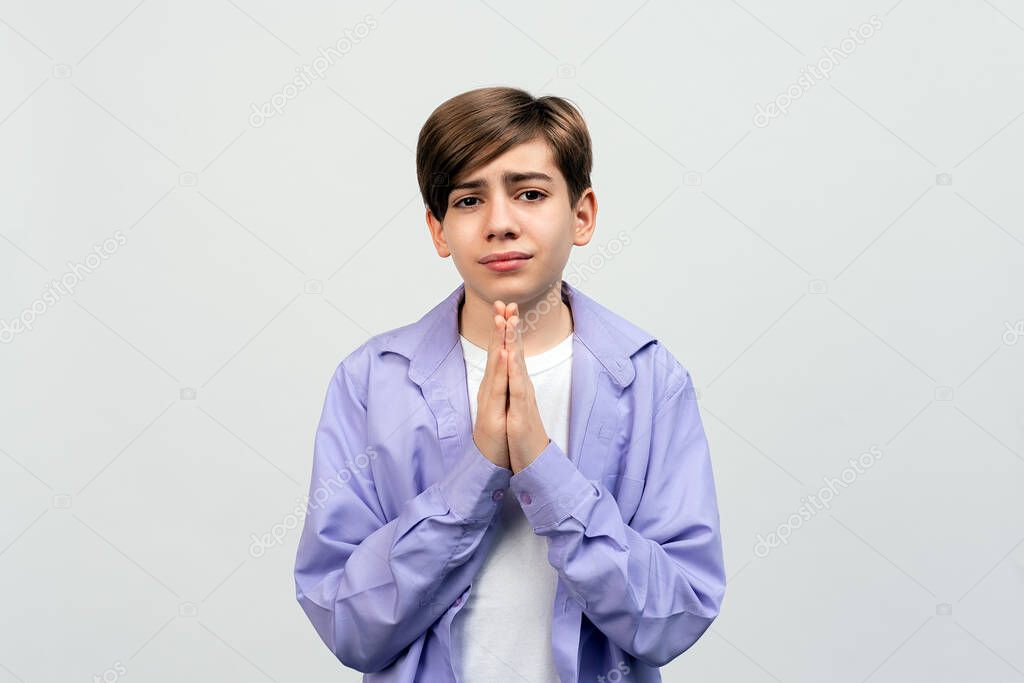 optimistic and hopeful teen boy praying with hands held together, waiting for results, awaiting miracle, praying with happy positive emotion, standing against light gray background