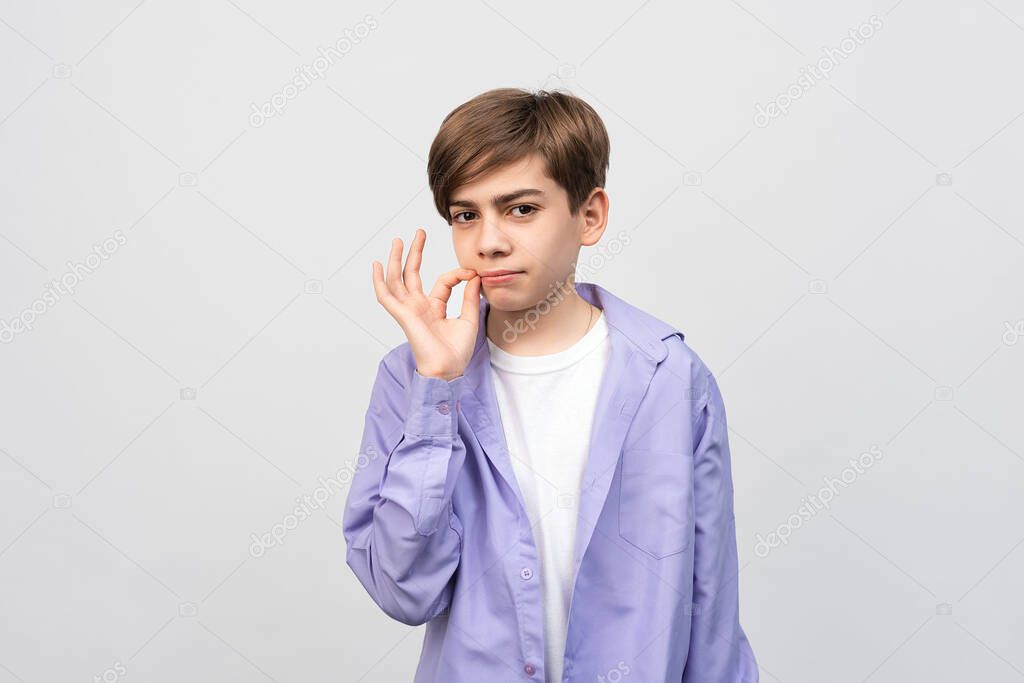Shh its secret. Portrait of serious boy 12-14 years old showing zip gesture as if shutting mouth on key, promises to keep secret, wearing casual clothes, gray background, isolated