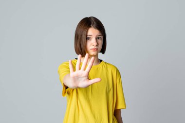 Ciddi bir genç kızın portresi, sarı günlük tişörtlü, duruş, yasaklama, blok hareketi gösteren, açık gri arka planda duran. Jesti durduran genç kız eli. Hayır, duruyoruz.