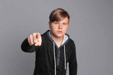 Ciddi bir genç çocuk kameraya kızgın ve sinirli bir şekilde seni işaret ediyor. Gri arka planda dikiliyor, günlük giysiler giyiyor.