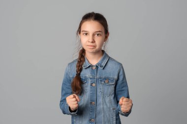 Genç kız kameraya negatif yüz ifadesiyle bakıyor yumruklarını öfkeyle kaldırıyor, gri arka planda izole edilmiş, kot ceket giyiyor. Etkileyici yüz ifadeleri. Öfke ve agresif kavram