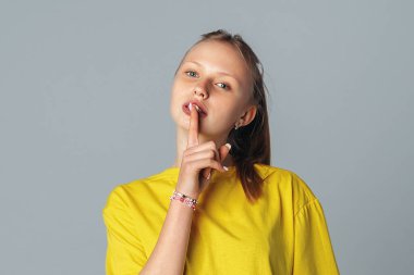 Güzel genç kız işaret dilini işaret parmağıyla dokunmaktan vazgeçmen için işaret dilini gösteriyor açık gri arka planda izole edilmiş, gündelik sarı tişört giyiyor. Etkileyici yüz ifadeleri.