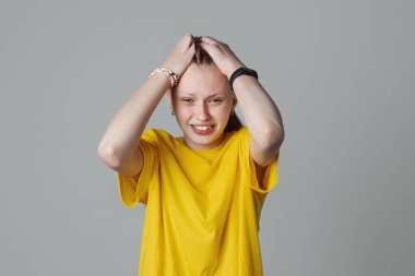 Baş ağrısı, panik ve depresyondan muzdarip, açık gri arka planda izole edilmiş, sarı tişörtlü, iki eliyle başını sımsıkı tutan genç bir kız.