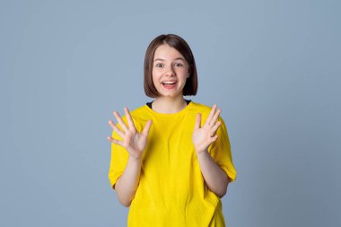 Sürpriz yüz ifadesi olan, sarı günlük tişört giymiş açık gri arka planda duran neşeli genç kız. Sürpriz kavramı