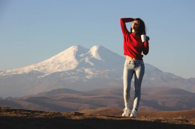 Kırmızı kazaklı güzel bir kadın dağların bulanık arka planında duruyor elinde çay fincanıyla güneşin ilk ışıklarının tadını çıkarıyor..