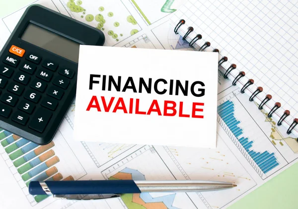 Business card with text Financing Available laying on financial graphs with notepad calculator and pen