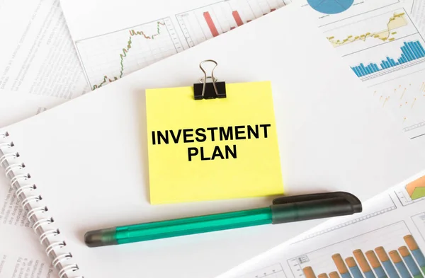 A yellow sticker with text Investment Plan is in a Notepad with a green pen financial charts and documents. Business and financial concept