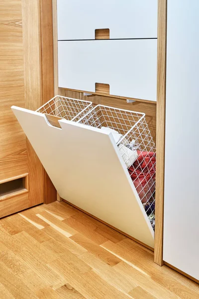 Modern wardrobe with opened metal mesh laundry basket. Wooden wardrobe with flat finger pull wardrobe doors. Oak veneered plywood cabinets with light gray painted cabinet doors