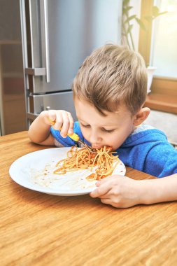 Büyük gözlü sevimli çocuk modern mutfakta metal buzdolabının karşısında oturan sarı çatallı, lezzetli makarna yiyor.