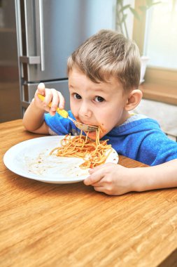 Büyük gözlü sevimli çocuk modern mutfakta metal buzdolabının karşısında oturan sarı çatallı, lezzetli makarna yiyor.