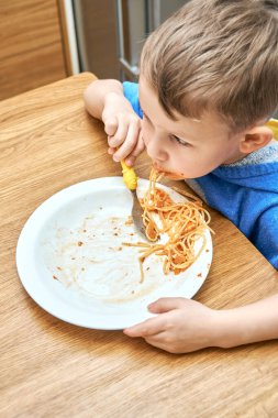Büyük gözlü sevimli çocuk modern mutfakta metal buzdolabının karşısında oturan sarı çatallı, lezzetli makarna yiyor.
