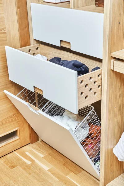 Modern wardrobe with opened metal mesh laundry basket and wooden drawers. Wooden wardrobe with flat finger pull wardrobe doors. Oak veneered plywood cabinets with light gray painted cabinet doors